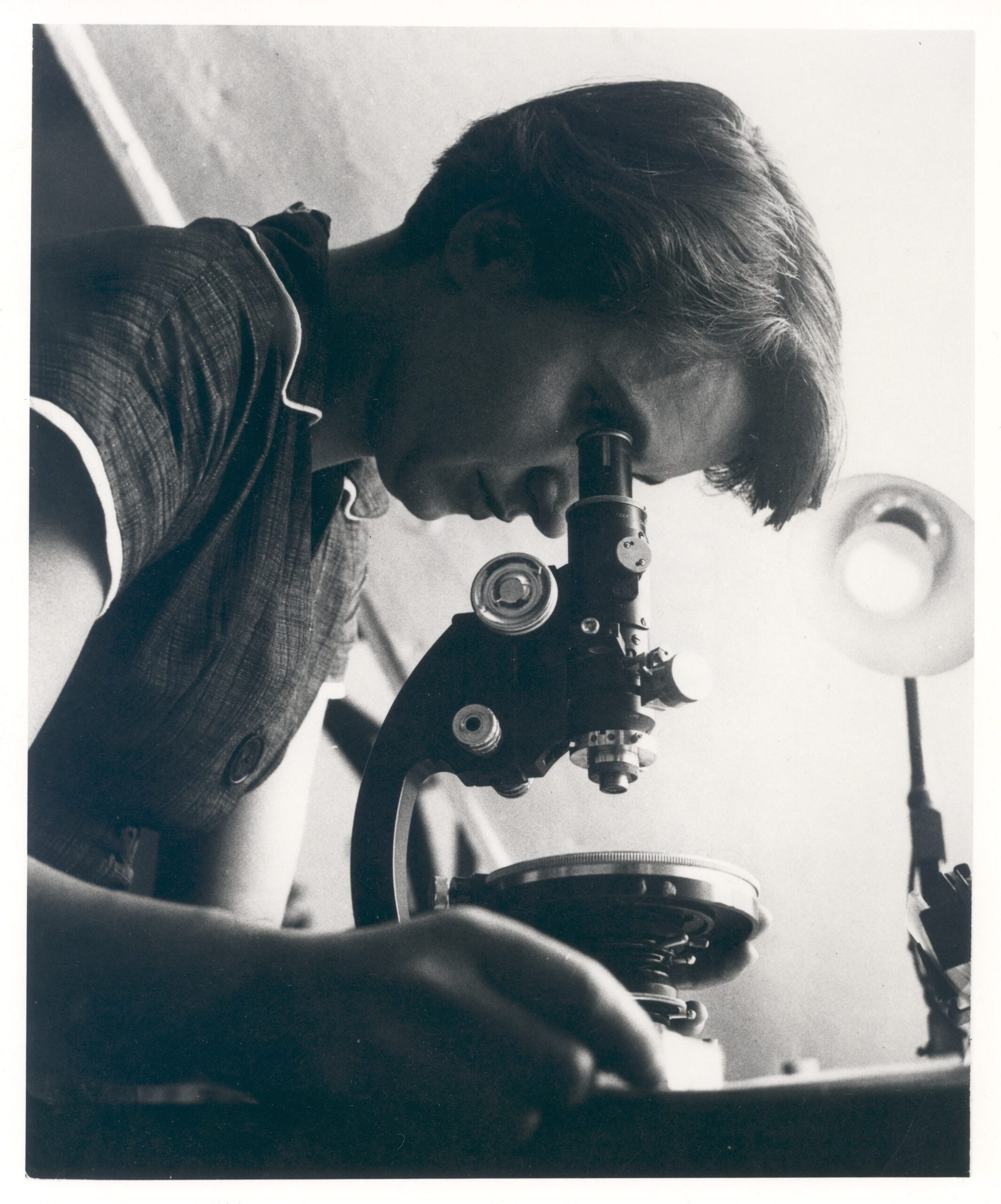 A black and white photograph of Rosalind Franklin looking into a microscope